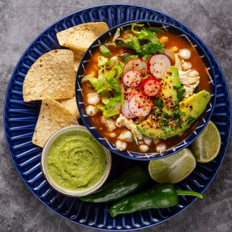 Pozole soup with avocado, radishes, and lettuce, served with tortilla chips, guacamole, and limes.